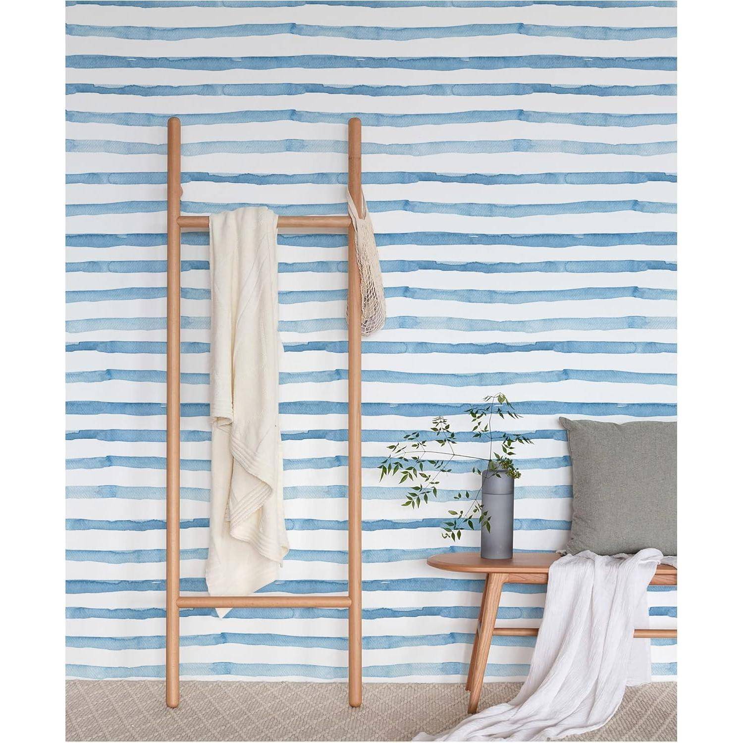 Eine Holzleiter lehnt mit zwei Handtüchern an einer blau-weiß gestreiften Wand. Eine Holzbank mit einem Kissen und einer Vase mit Zweigen steht in der Nähe.