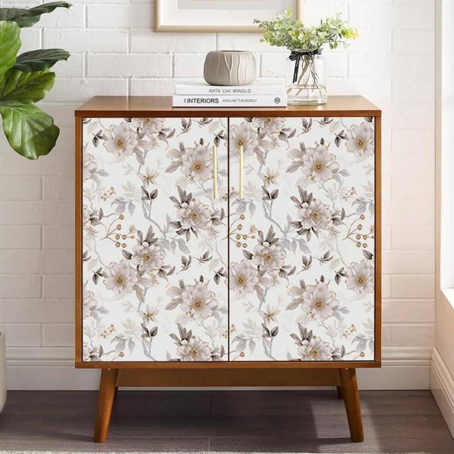 Ein Holzschrank mit blumenmusterierten Türen steht an einer weißen Ziegelwand. Eine Pflanze, ein Buch und eine Vase befinden sich auf der Oberfläche des Schranks.
