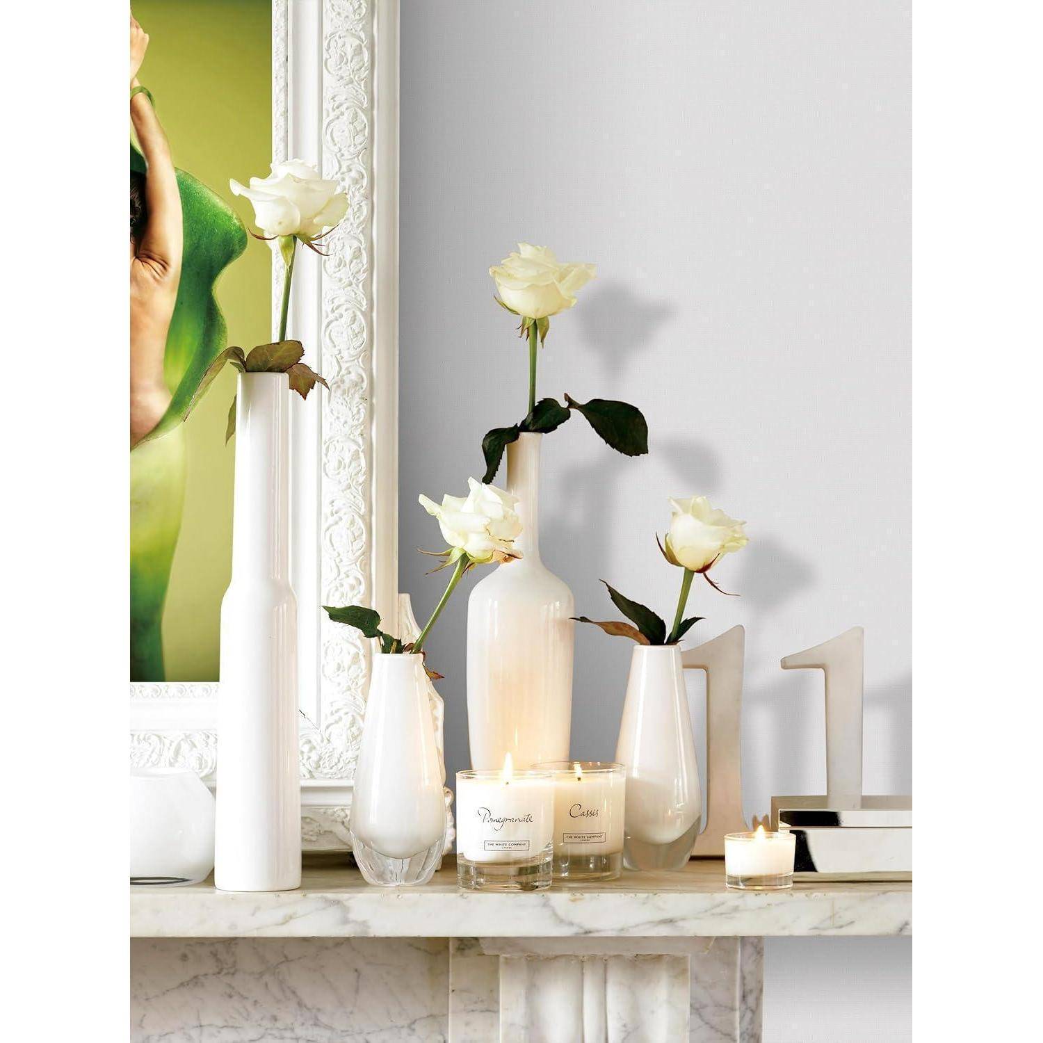 Weiße Rosen in eleganten Vasen und brennende Kerzen sind auf einem Marmorkamin angeordnet und schaffen eine ruhige und dekorative Anordnung.