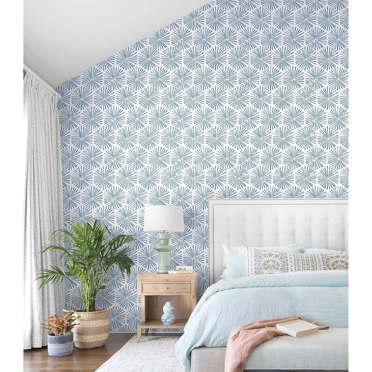 Schlafzimmer mit einem Bett, das mit pastellfarbener Bettwäsche bezogen ist, einem Holznachttisch mit einer Lampe und Topfpflanzen. Blaue gemusterte Tapete an der Akzentwand.