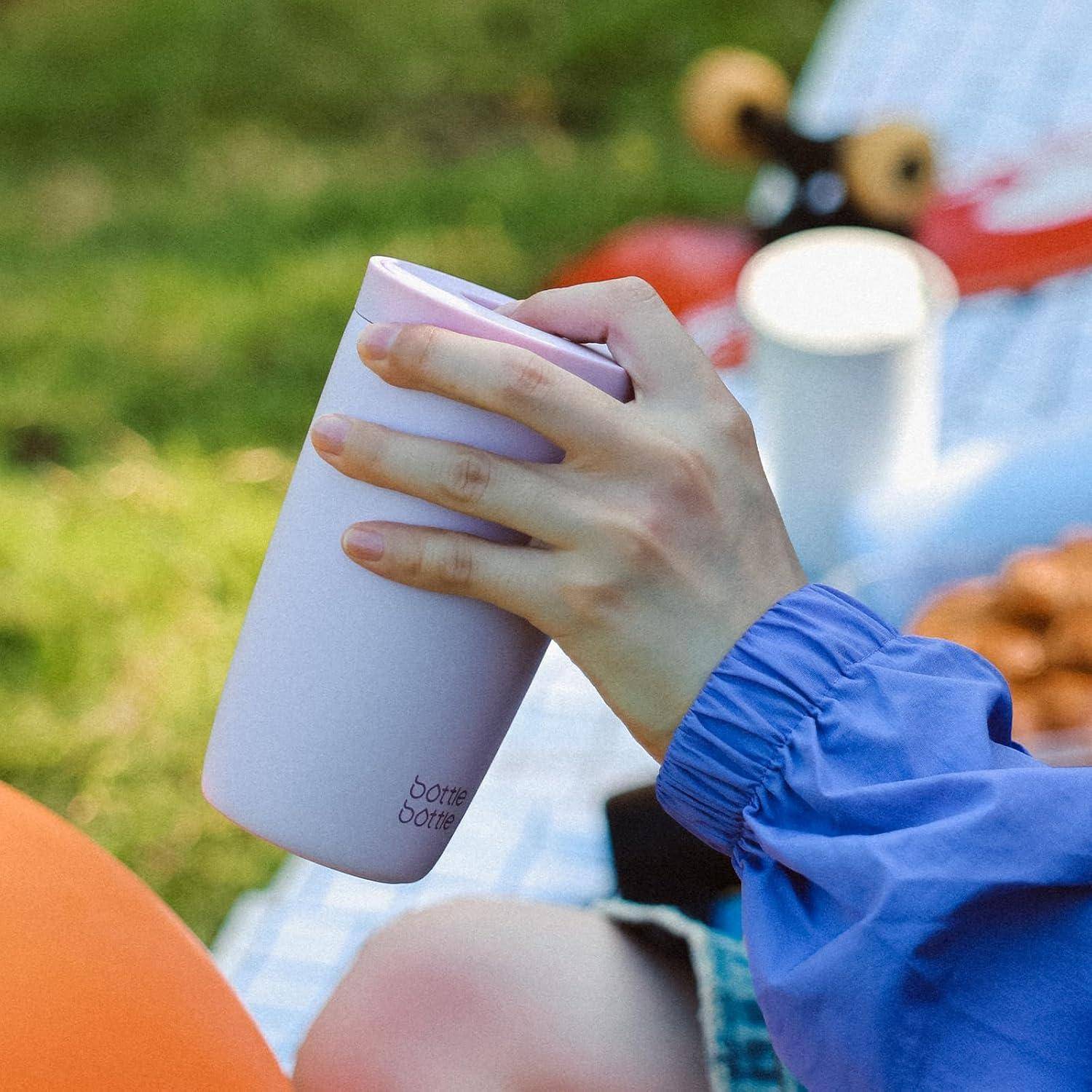 Eine Hand, die eine mit Lavendel isolierte Flasche auf einer Picknickdecke hält, umgeben von unscharfen Picknickgegenständen und einem grasigen Hintergrund.