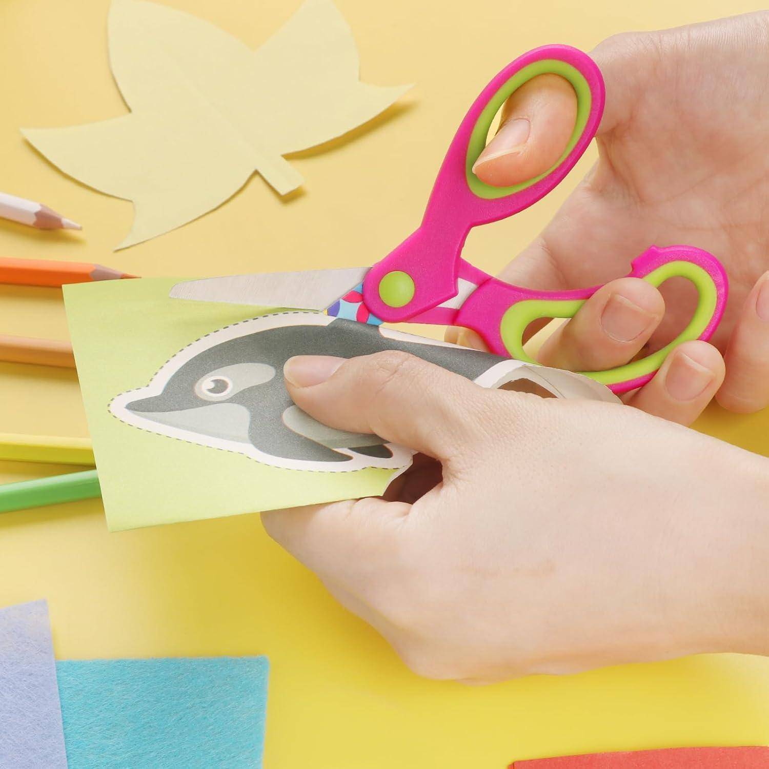 Eine Person schneidet einen gedruckten Pinguin mit rosa Schere auf grünem Papier. Bunte Papiere und Buntstifte sind verstreut umher.
