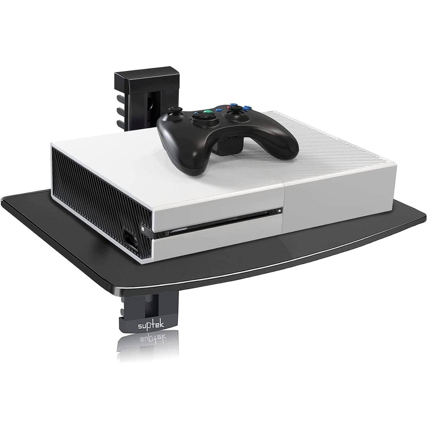 Eine weiße Spielkonsole mit einem schwarzen Controller darauf, die auf einem an der Wand befestigten schwarzen Regal platziert ist.