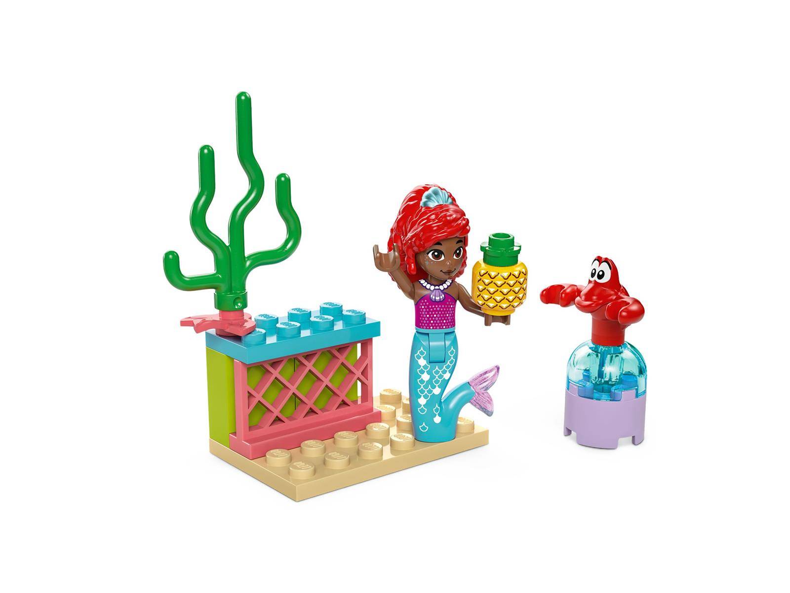 LEGO-artige Ariel-Minipuppe neben einer Ananas auf einer kleinen Strandszene mit einer grünen korallenähnlichen Pflanze und einer roten Krabbenfigur auf einem Ständer.