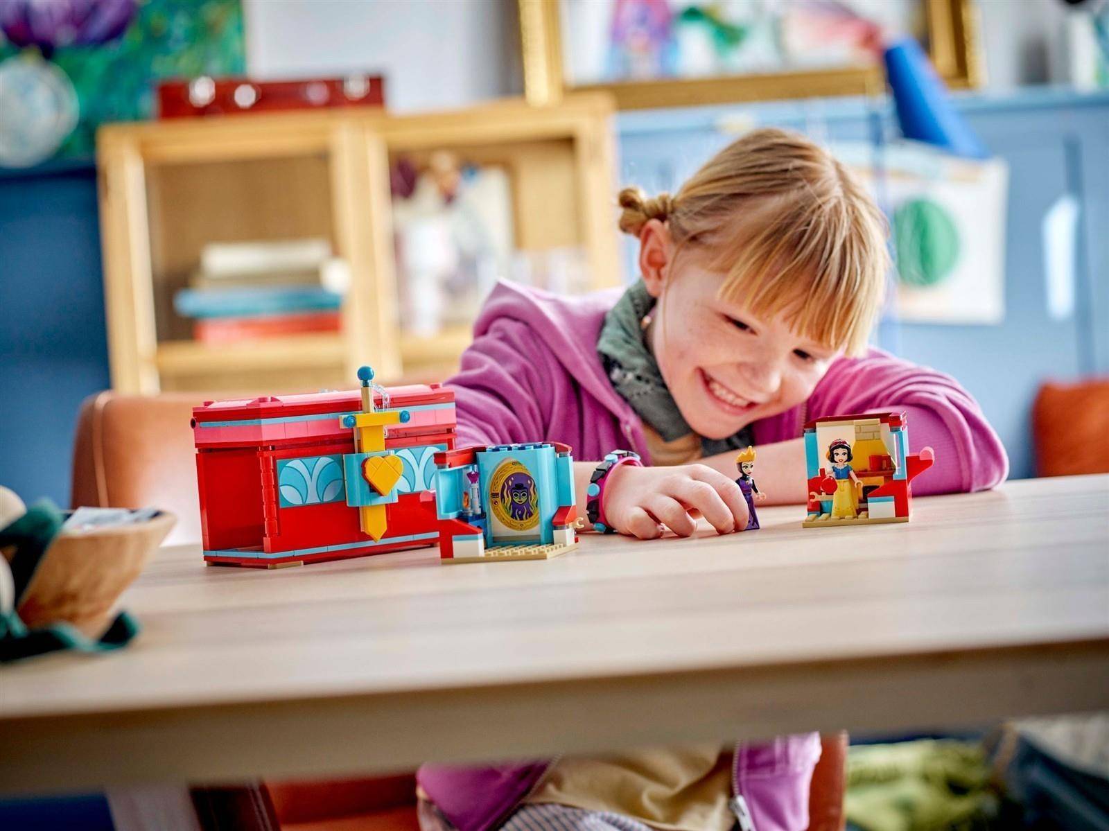 Lächelndes Kind beugt sich über einen Tisch und spielt mit farbenfrohen LEGO-ähnlichen Spielzeuggebäuden und Figuren in einem Klassenzimmer oder Spielzimmer.