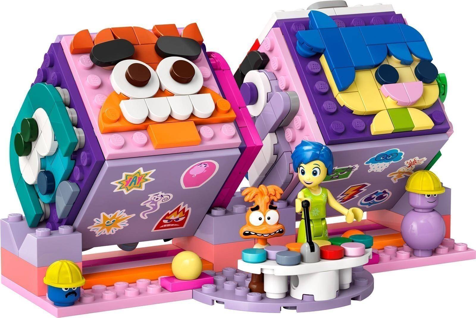 Zwei bunte LEGO-ähnliche Monsterboxen mit Gesichtern, eine orange und eine blau, stehen auf einem Spielset mit einer Jungen-Minifigur und kleinen Zubehörfiguren davor.
