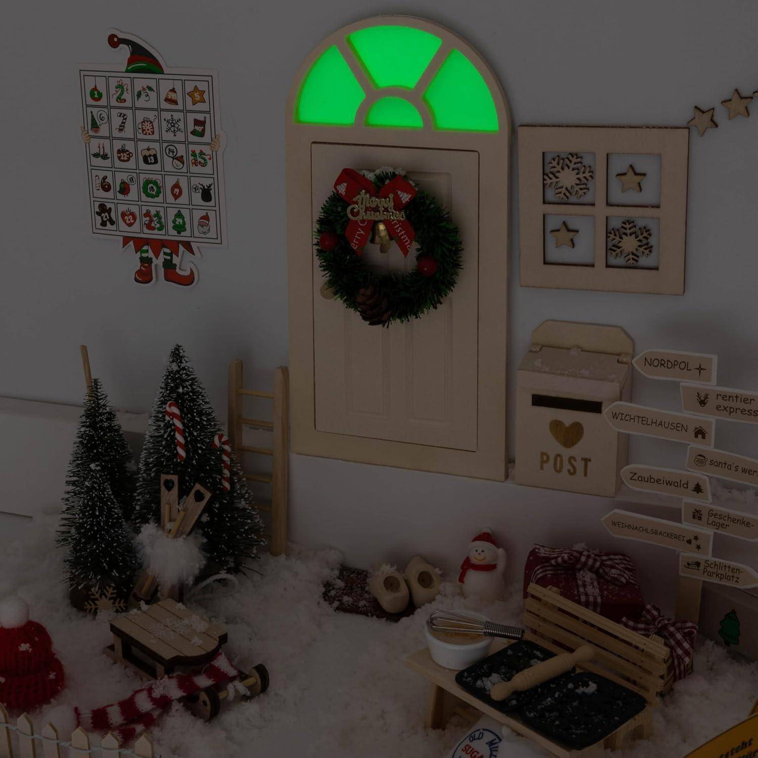 Eine Weihnachts-Miniaturszene zeigt eine kleine dekorierte Tür mit einem grün leuchtenden Fenster, einem Kranz und winzigen Weihnachtsornamente.