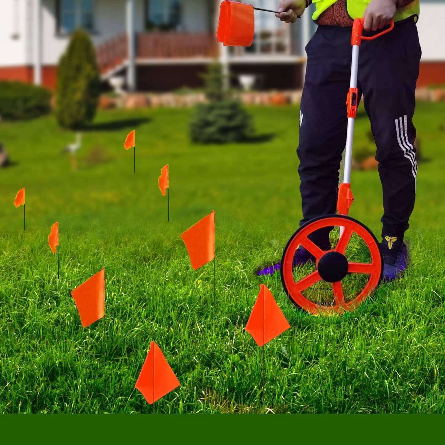 Eine Person steht mit einem Messrad auf einer grasbewachsenen Rasenfläche, wobei mehrere orangefarbene Flaggen Punkte auf dem Boden markieren.