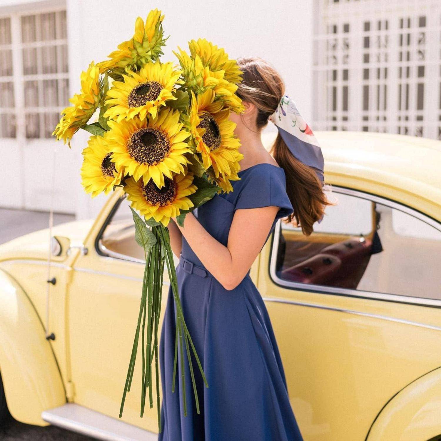 Frau in einem blauen Kleid, die einen großen Strauß Sonnenblumen hält, steht neben einem gelben Oldtimer, wobei ihr Kopf abgewandt ist.