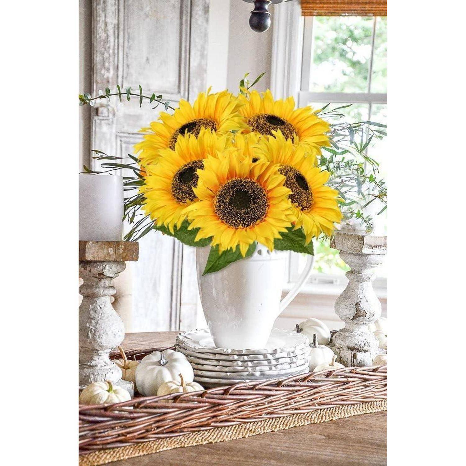 Ein weißer Krug enthält strahlend gelbe Sonnenblumen auf einem rustikalen Holztisch, umgeben von weißen Kürbissen und Kerzen, neben einem Fenster.
