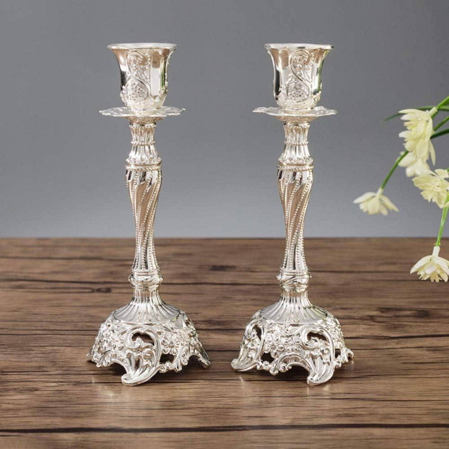 Zwei kunstvoll gearbeitete silberne Kerzenhalter mit filigranen Details stehen auf einer Holzoberfläche. Rechts ist eine kleine weiße Blume zu sehen.