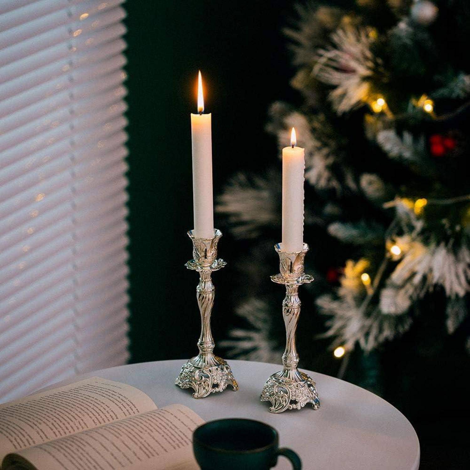 Zwei angezündete Kerzen auf kunstvollen Haltern stehen auf einem Tisch mit einem aufgeschlagenen Buch und einer Tasse. Im Hintergrund befindet sich ein unscharfer Weihnachtsbaum mit Lichtern.