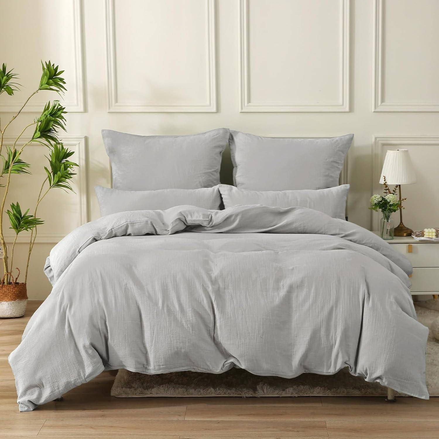 Ein ordentlich gemachtes Bett mit hellgrauer Bettwäsche, zwei großen Kissen und einer passenden Bettdecke. Ein kleiner Nachttisch mit einer Lampe und einer Pflanze steht daneben.