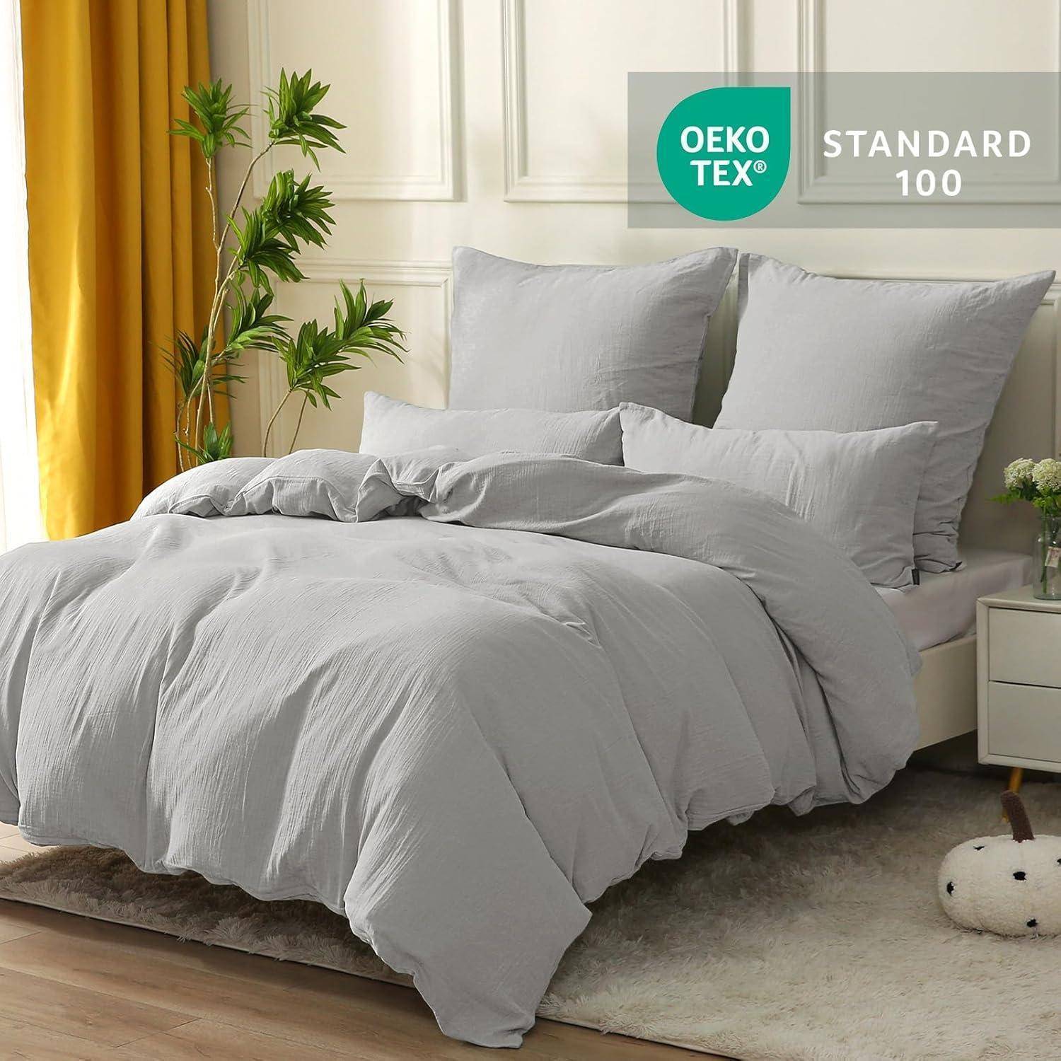 Ein ordentlich gemachtes Bett mit hellgrauer Bettwäsche, flankiert von einer Pflanze und einer Lampe. Zertifizierungslabel 'OEKO-TEX STANDARD 100' in der Ecke sichtbar.