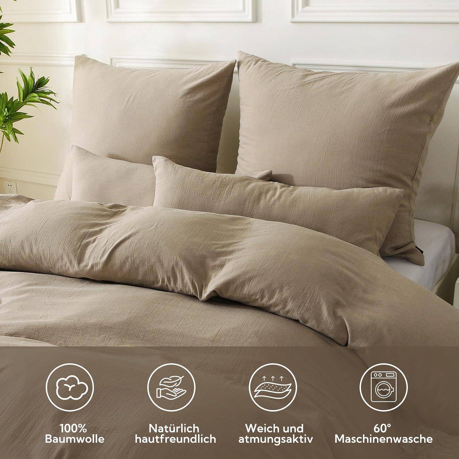 Ein ordentlich gemachtes Bett mit taupefarbener Bettwäsche umfasst zwei große Kissen, ein mittleres Kissen und eine Bettdecke. Die Icons darunter heben Merkmale hervor: '100% Baumwolle', 'Natürlich hautfreundlich', 'Weich und atmungsaktiv', '60° maschinenwaschbar'.