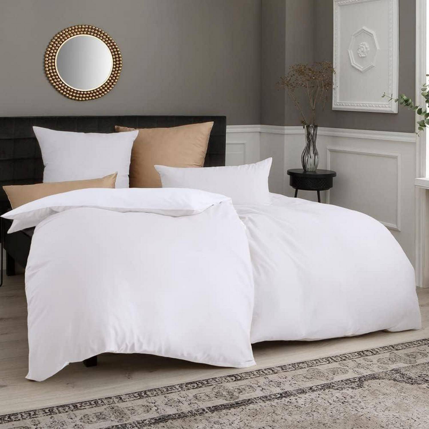 Ein modernes Schlafzimmer mit einem ordentlich gemachten Bett, bezogen mit weißen Bettwäsche, zwei Kissen und einem dunkelgrauen gepolsterten Kopfteil. An der Wand hängen ein rundes Spiegel und gerahmte Kunstwerke.