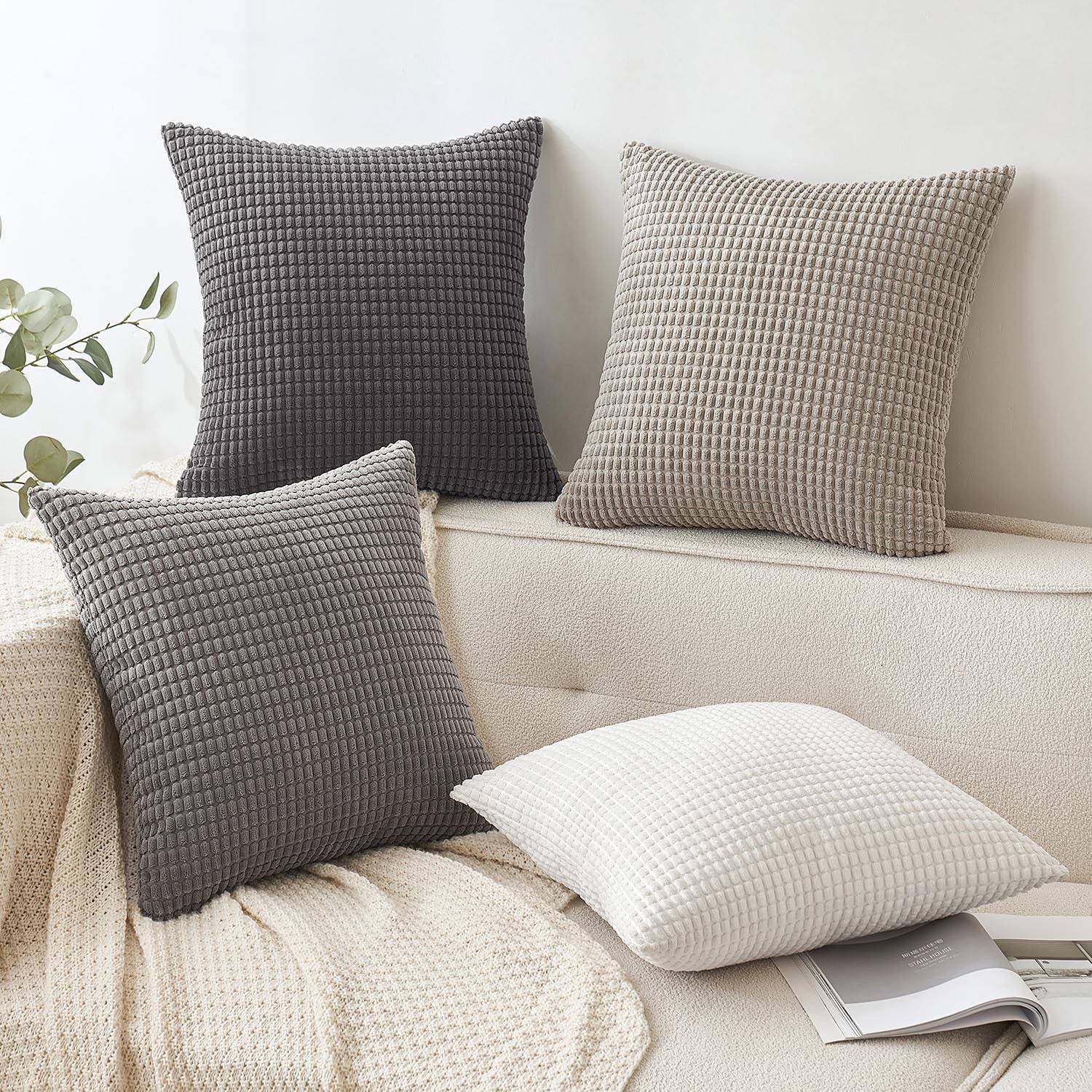Vier Kissen in verschiedenen Schattierungen von Grau und Weiß sind auf einem Sofa angeordnet, daneben liegt eine strukturierte Decke, und im Hintergrund befindet sich eine Pflanze.