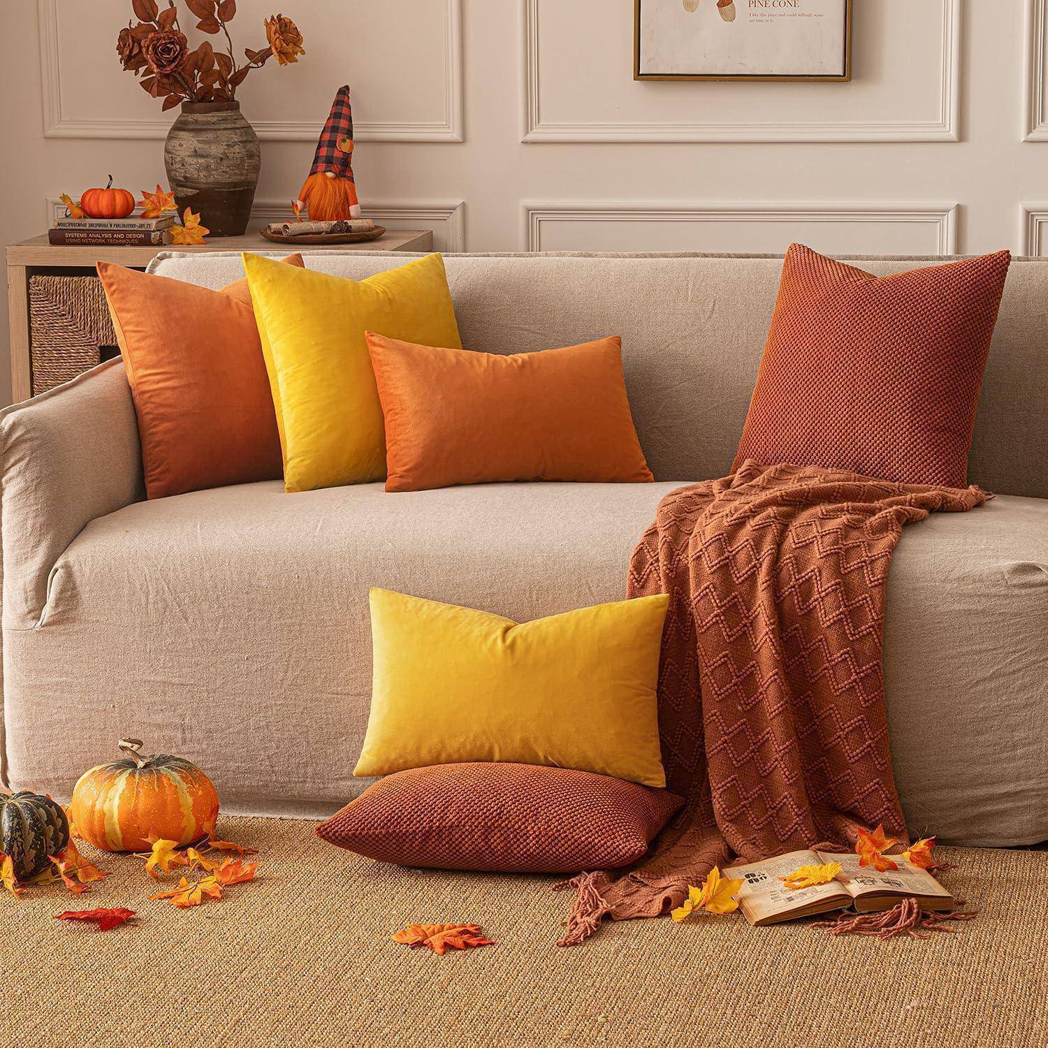 Ein gemütliches Wohnzimmer mit einem beigen Sofa, geschmückt mit orange- und gelben Kissen. Herbstblätter und Kürbisse verleihen eine festliche, jahreszeitliche Note.