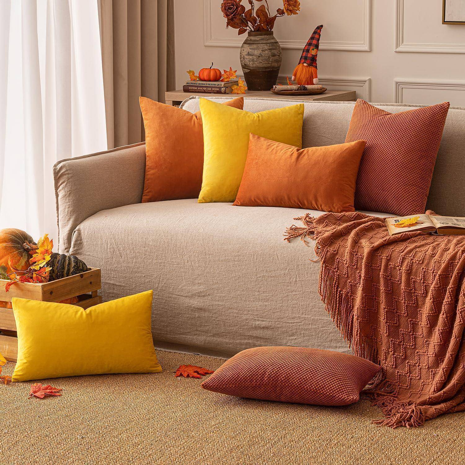 Ein gemütliches Wohnzimmer mit einem beigen Sofa, geschmückt mit orange- und gelben Kissen. Eine Decke fällt über das Sofa, mit Herbstdekoration in der Nähe.