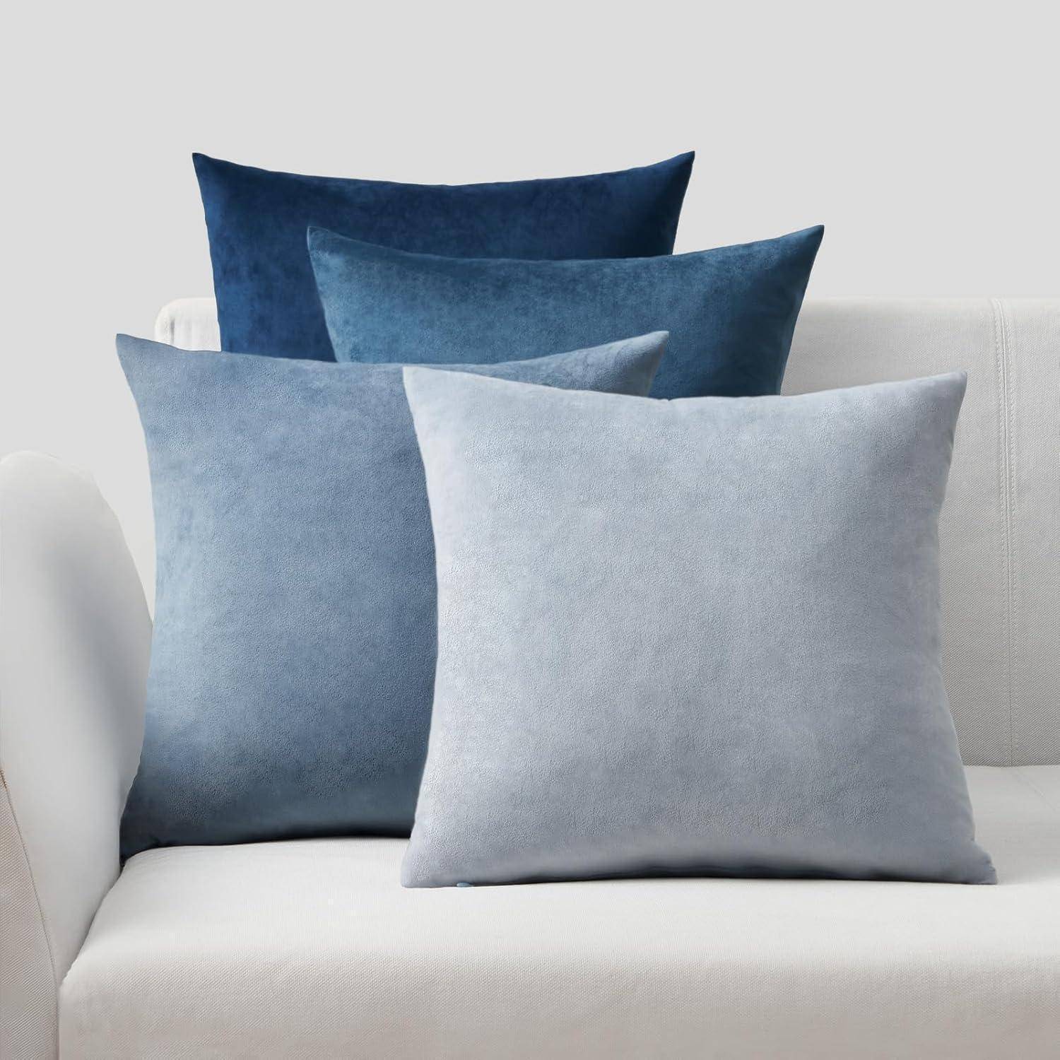 Vier quadratische Samtkissen in Blau- und Grautönen sind auf einem weißen Sofa gestapelt.