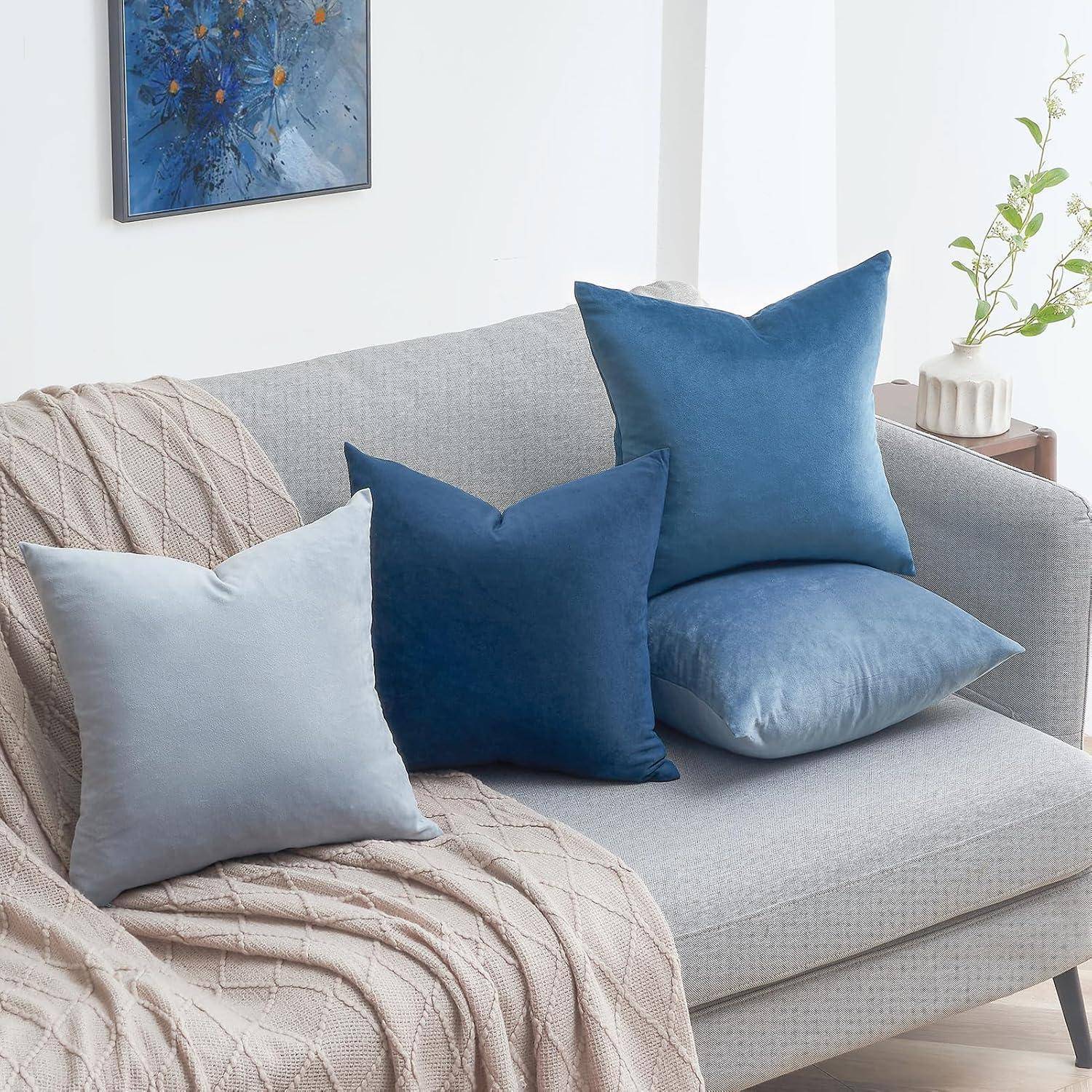 Ein hellgrauer Sofa, geschmückt mit blauen und hellgrauen Kissen, überzogen mit einer beigen gestrickten Decke. Ein Blumengemälde hängt im Hintergrund.