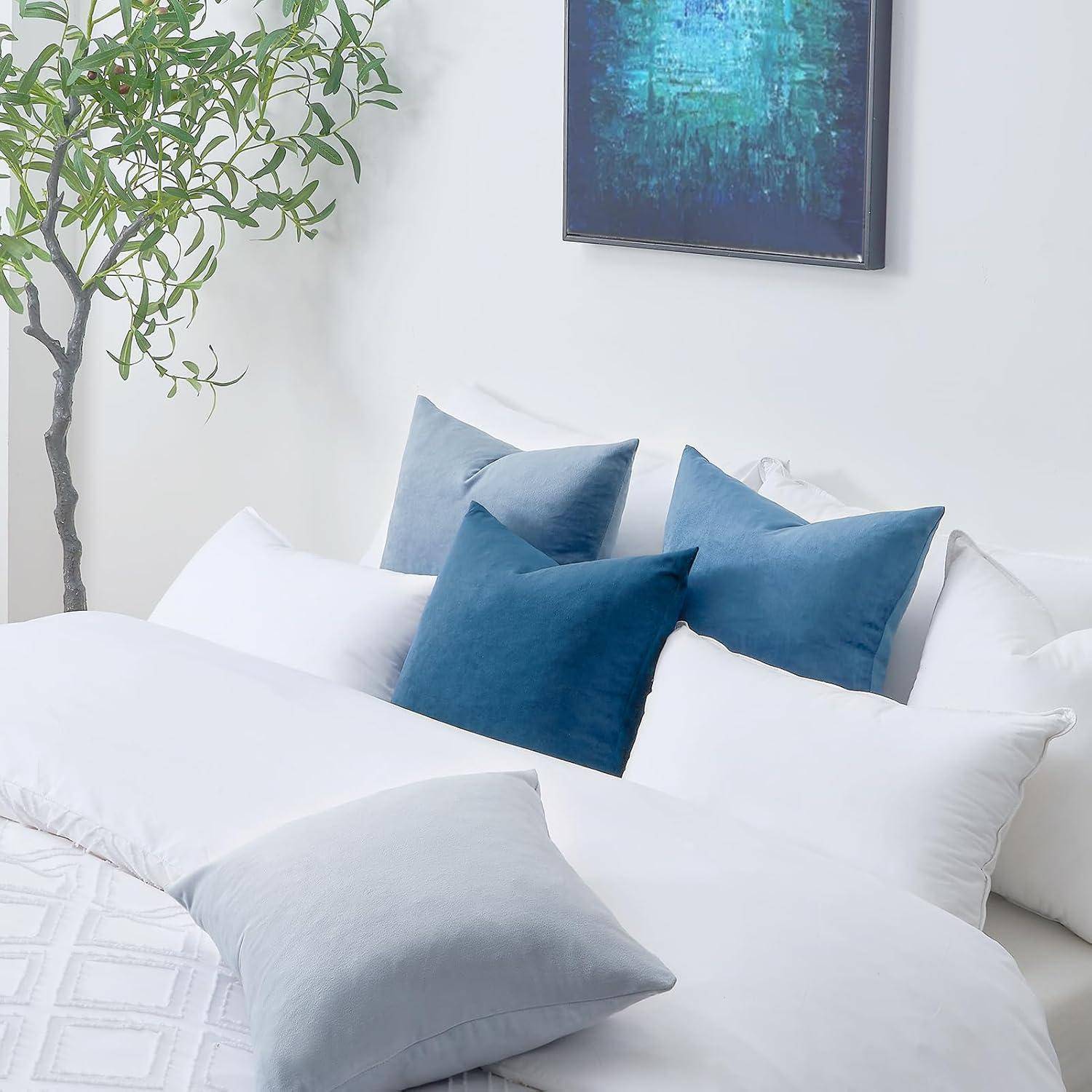 Eine helle Schlafzimmerszene mit einem sorgfältig gemachten weißen Bett, das blaue und weiße Kissen trägt, eine blattreiche Zimmerpflanze und ein blaues abstraktes Gemälde an der Wand.