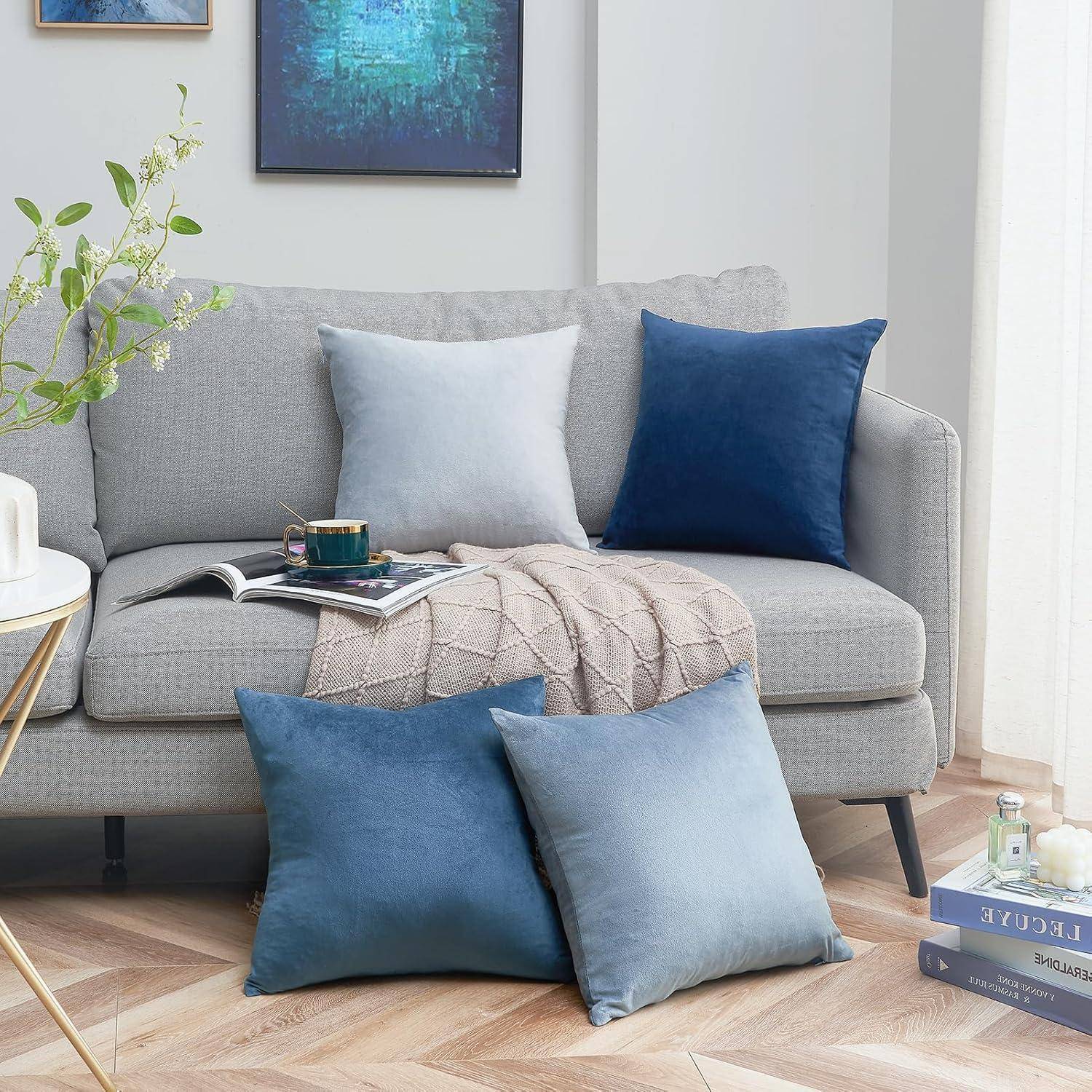 Ein moderner Wohnbereich mit einem hellgrauen Sofa, geschmückt mit blauen und grauen Kissen, einer gemütlichen Decke und einer kleinen Pflanze auf einem runden Tisch.