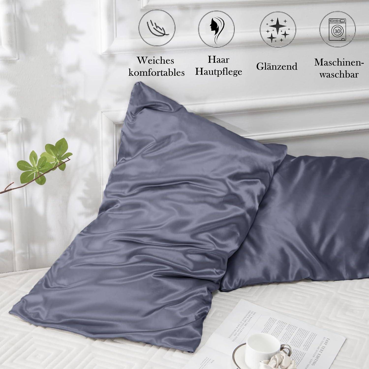 Zwei graue Seidenkissenbezüge auf einem Bett, mit Symbolen darüber, die Weichheit, Haar- und Hautpflege sowie Maschinenwäsche anzeigen. In der Nähe befinden sich eine Tasse und ein Zweig.