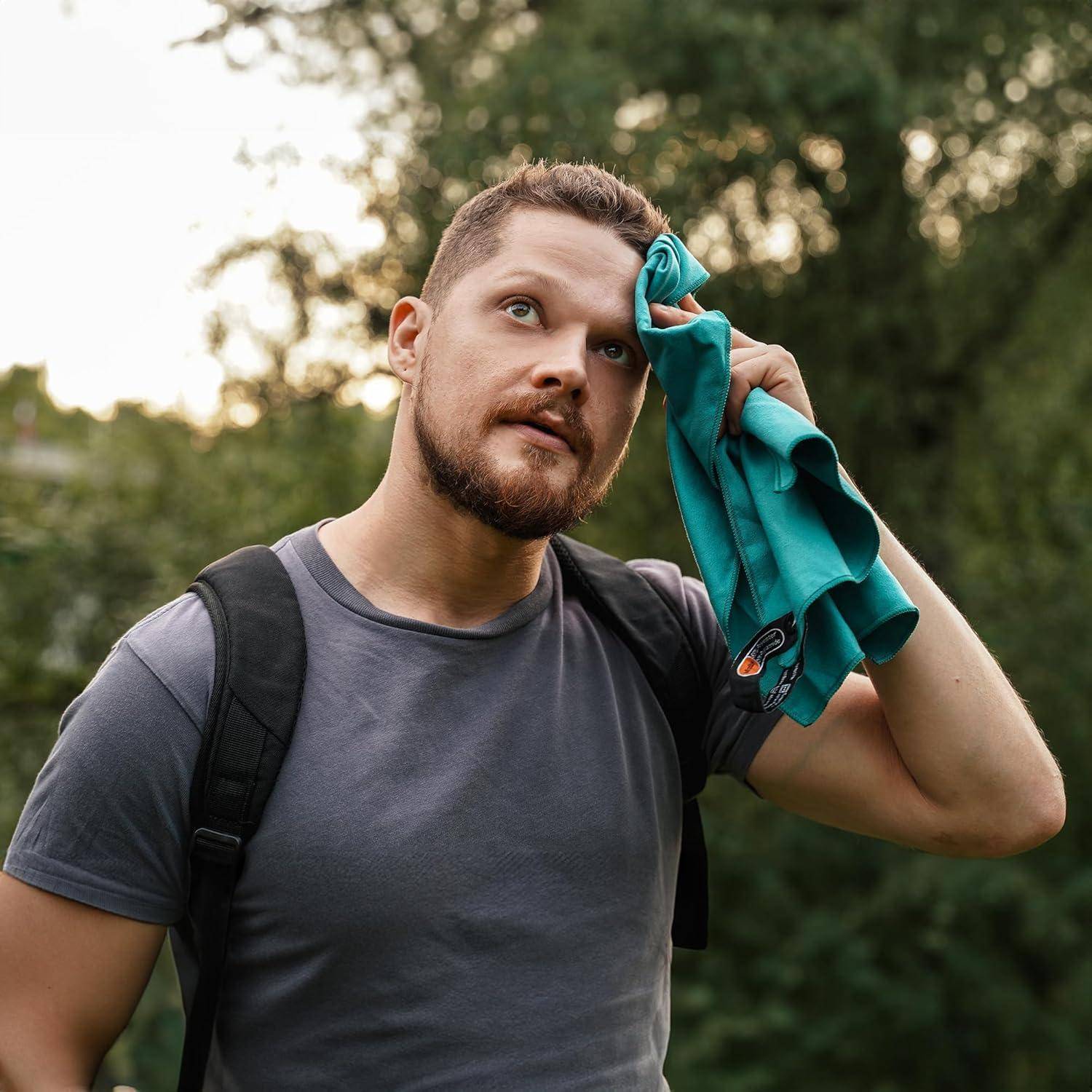 Ein Mann mit Bart, der ein graues T-Shirt und einen schwarzen Rucksack trägt, wischt sich mit einem türkisfarbenen Tuch die Stirn und steht im Freien neben Grünpflanzen.