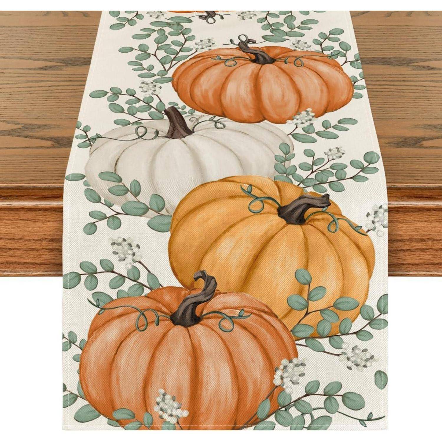 Tischläufer mit orangefarbenen und weißen Kürbissen und grünem Blätterdesign auf einem Holztisch.