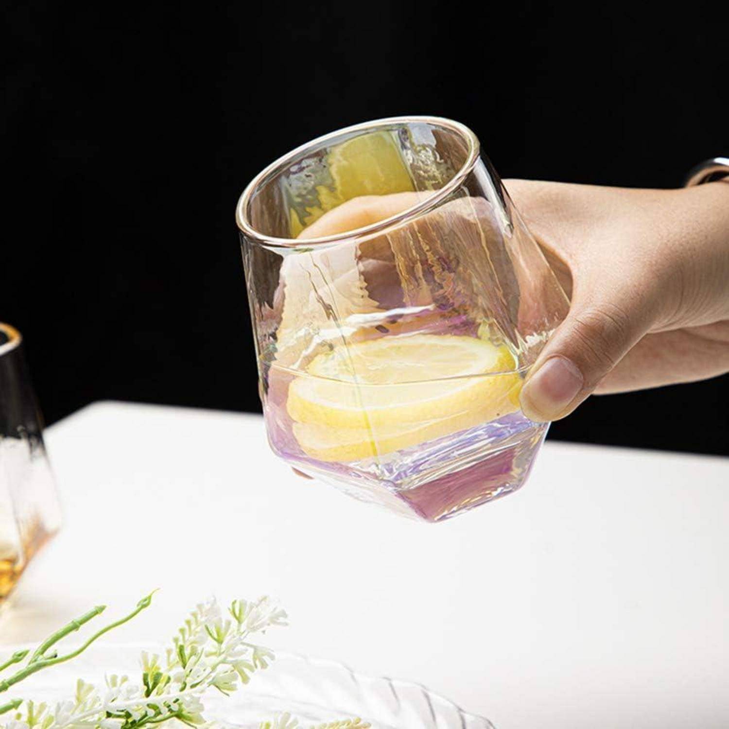 Eine Hand hält ein klares Glas mit einer Orangenscheibe und Flüssigkeit. In der Nähe vervollständigen ein Zweig Grün und ein Teller die Tischszene.