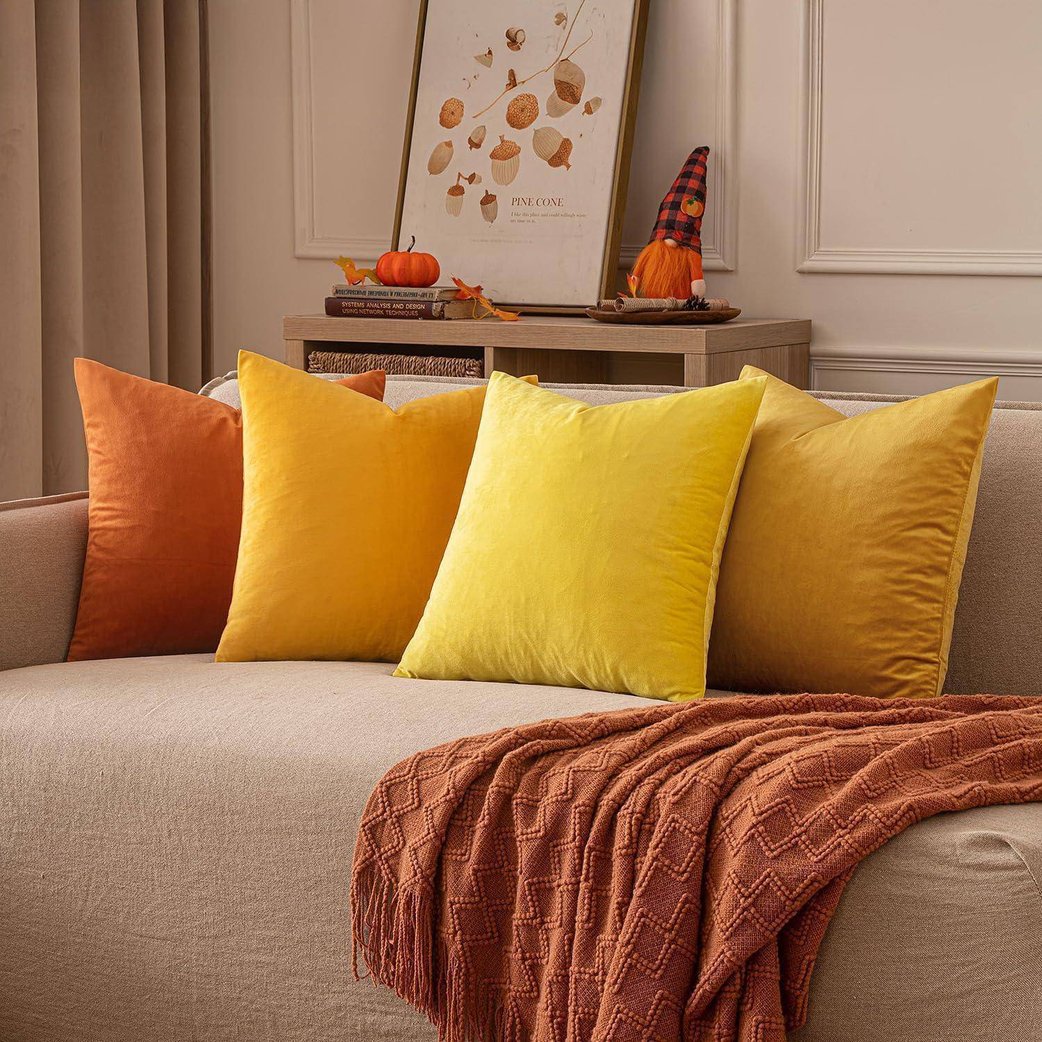 Ein beige Sofa mit orange und gelben Kissen und einer orangefarbenen Decke steht vor einer Wand mit herbstlicher Dekoration und gerahmten Kunstwerken.