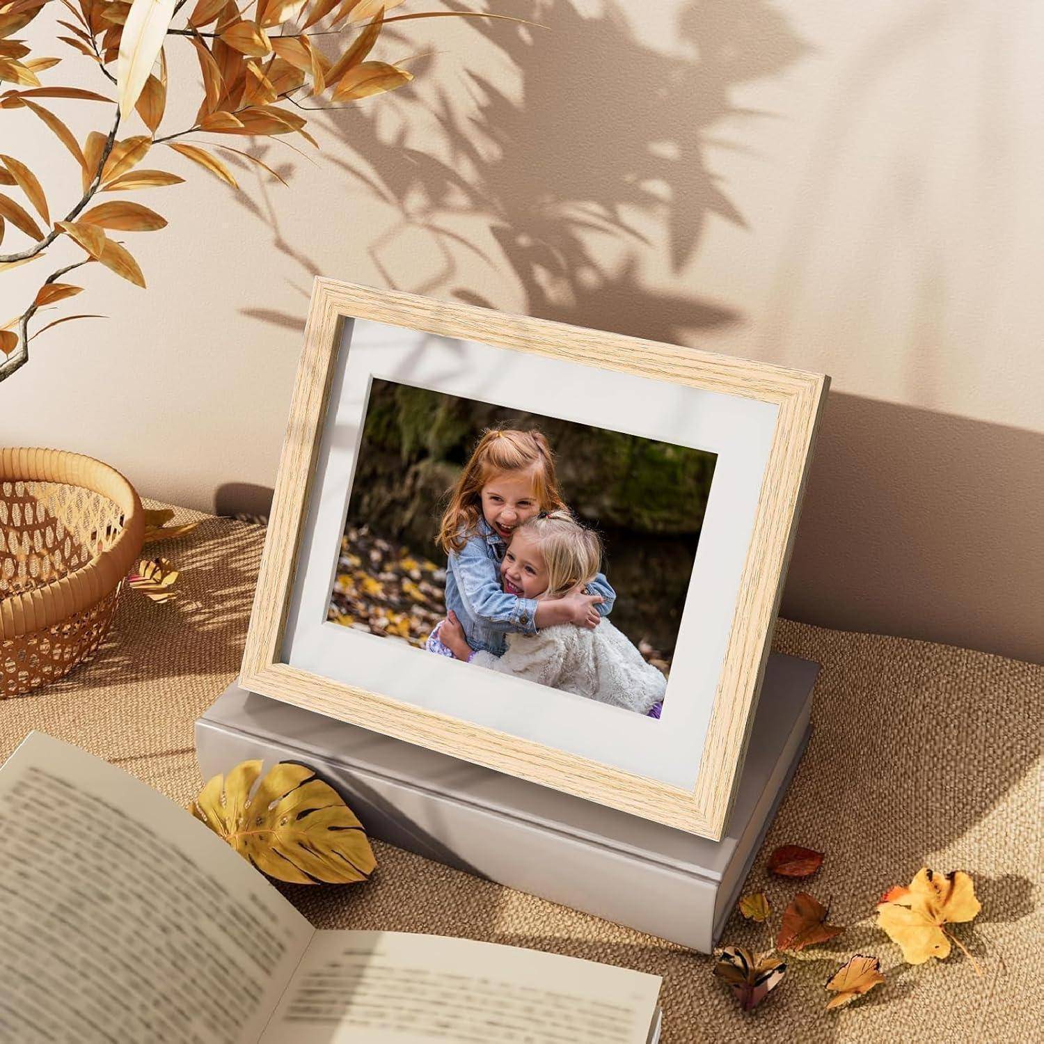 Ein Fotorahmen auf einem Tisch zeigt zwei lächelnde Kinder, die sich umarmen, umgeben von Herbstblättern. Die Umgebung umfasst einen Weidenkorb und ein aufgeschlagenes Buch.