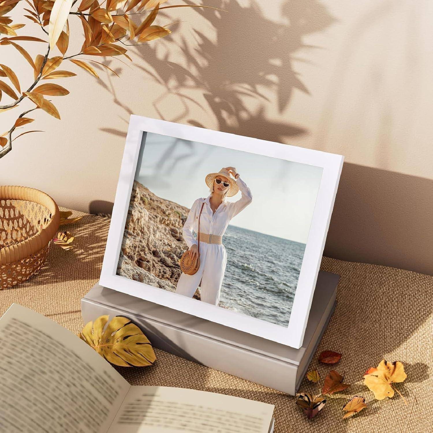 Ein gerahmtes Foto auf einem Tisch zeigt eine Person am Strand mit Sonnenhut, in Weiß gekleidet, neben einem Buch, einem Blatt und einer Vase, was eine warme, entspannte Atmosphäre schafft.