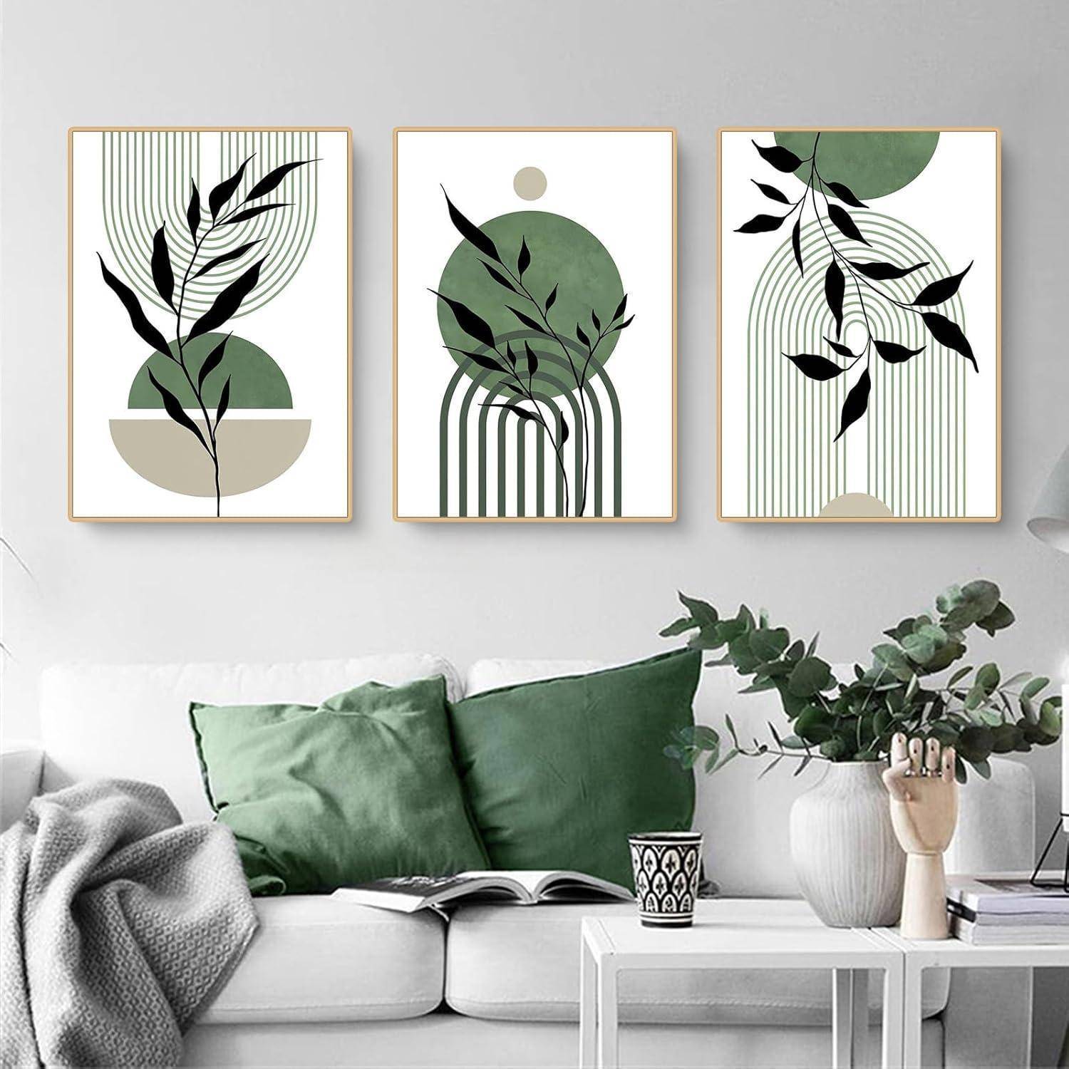 Drei abstrakte botanische Drucke in Grün- und Beigetönen hängen über einem weißen Sofa mit grünen Kissen und einem Buch auf der Armlehne in einem minimalistischen Wohnzimmer.
