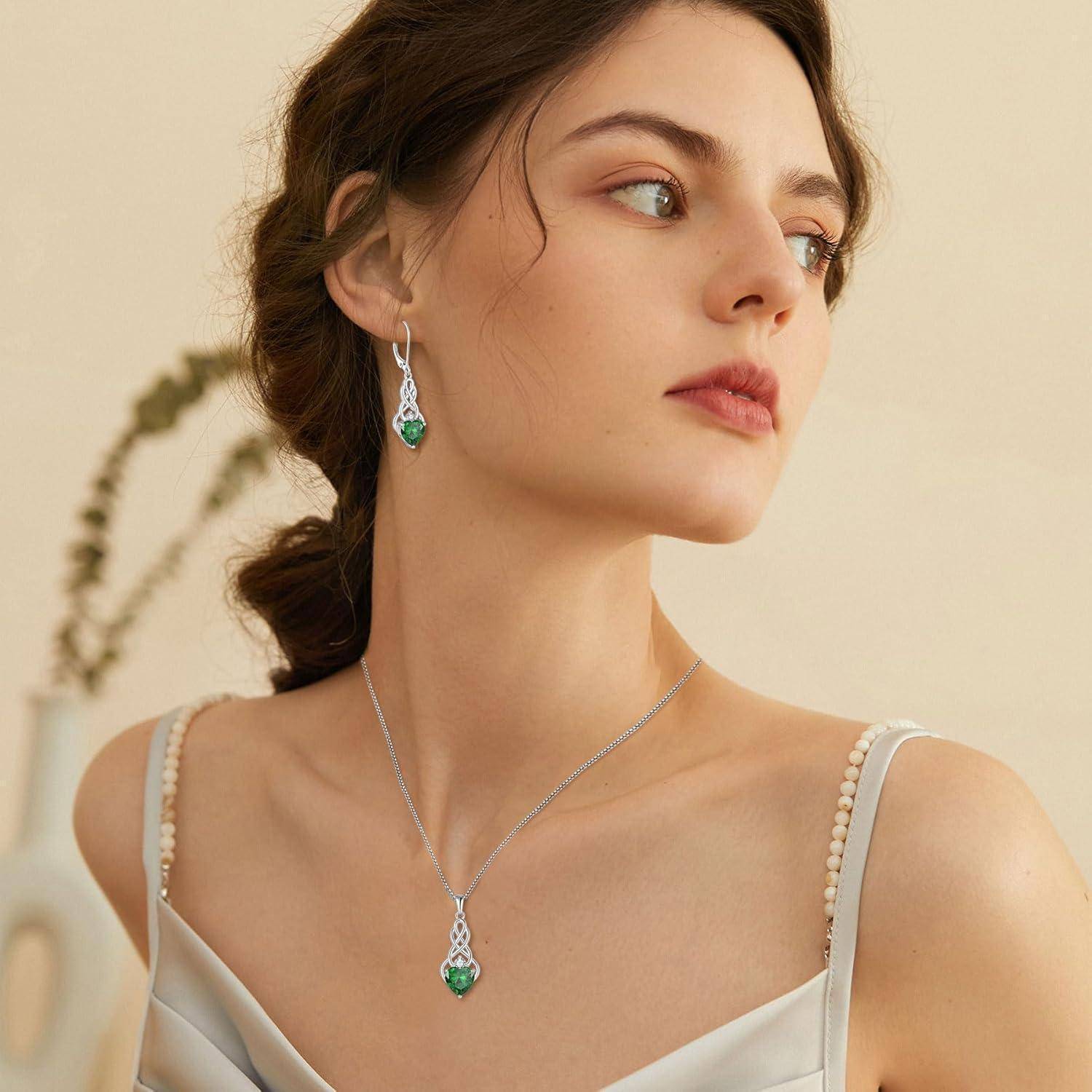 Eine Frau mit neutralem Gesichtsausdruck trägt eleganten grünen Edelschmuck und ein Seidenkleid, während sie in einer sanft beleuchteten Umgebung posiert.