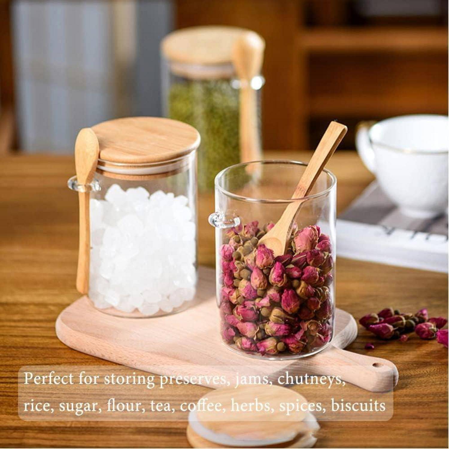 Zwei Glasgefäße mit Holzdeckeln und Schaufeln, eines gefüllt mit weißen Zuckerkristallen und das andere mit getrockneten Rosenblüten, auf einem Holzbrett.