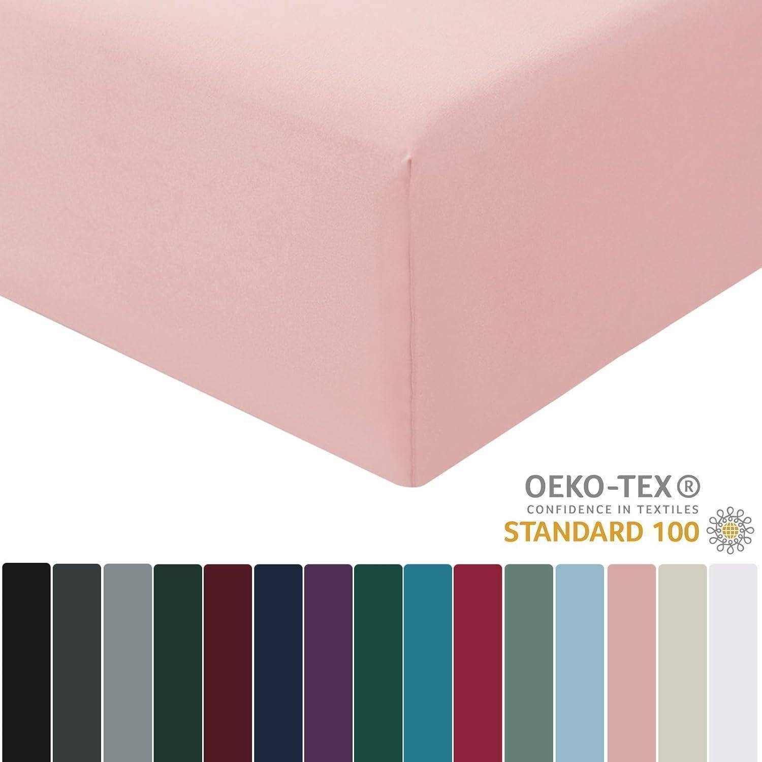 Eine pastellrosa Spannbettlaken-Ecke mit OEKO-TEX Standard 100 Etikett. Darunter Farbmuster in Schwarz, Grau, Grün, Blau und anderen Farben.