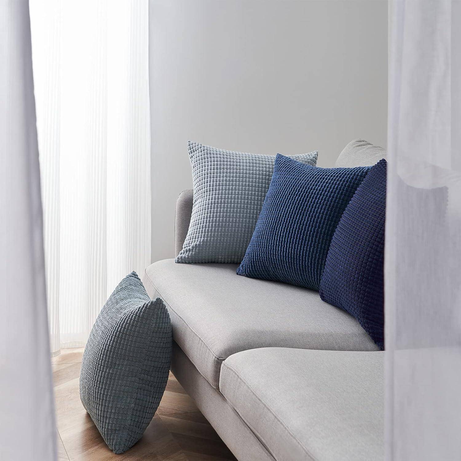 Eine hellgraue Couch mit vier strukturierten Kissen in Blau- und Grautönen steht neben einem transparenten, weißen Vorhang.