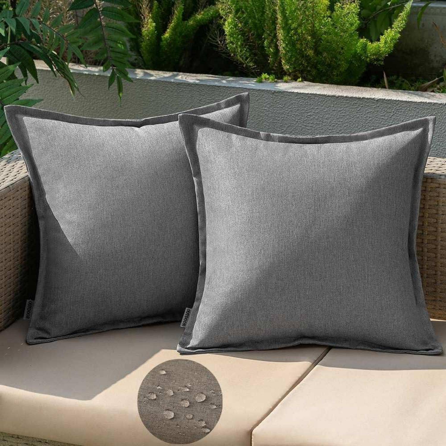 Zwei graue Kissen auf einem Rattansofa auf der Terrasse mit Pflanzen im Hintergrund.