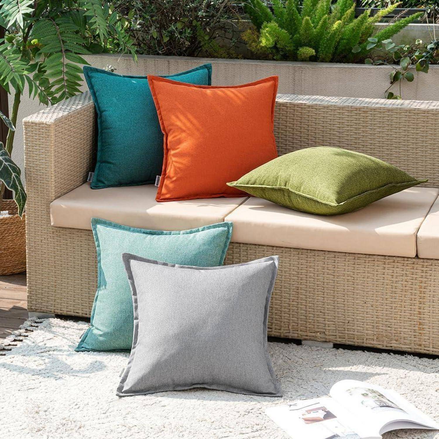 Eine beige Rattancouch mit bunten Kissen in Orange, Grün, Blau und Grau steht draußen auf einer Terrasse, umgeben von üppigen Blättern.