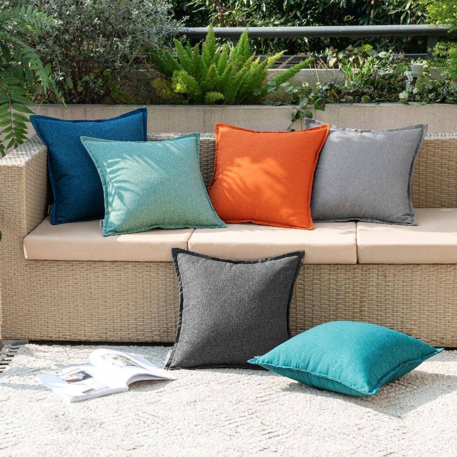 Ein hellbraunes Rattansofa mit bunten Kissen in Blau, Petrol, Orange und Grau auf einer Steinterrasse. Eine Zeitschrift und ein petrolblaues Kissen liegen davor.