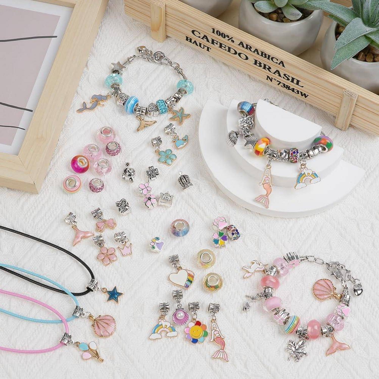 Eine Sammlung bunter Armbänder und Halsketten mit Anhängern, ausgelegt auf einem weißen Tisch. Eine kleine Holzkiste mit Sukkulenten befindet sich im Hintergrund.