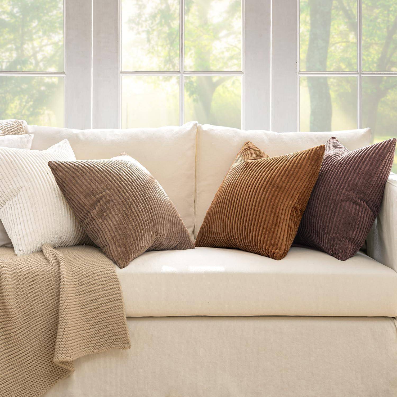 Ein beige Sofa mit verschiedenen Kissen, darunter strukturierte braune, gestreifte und einfarbig weiße Kissen, steht vor großen, sonnendurchfluteten Fenstern.