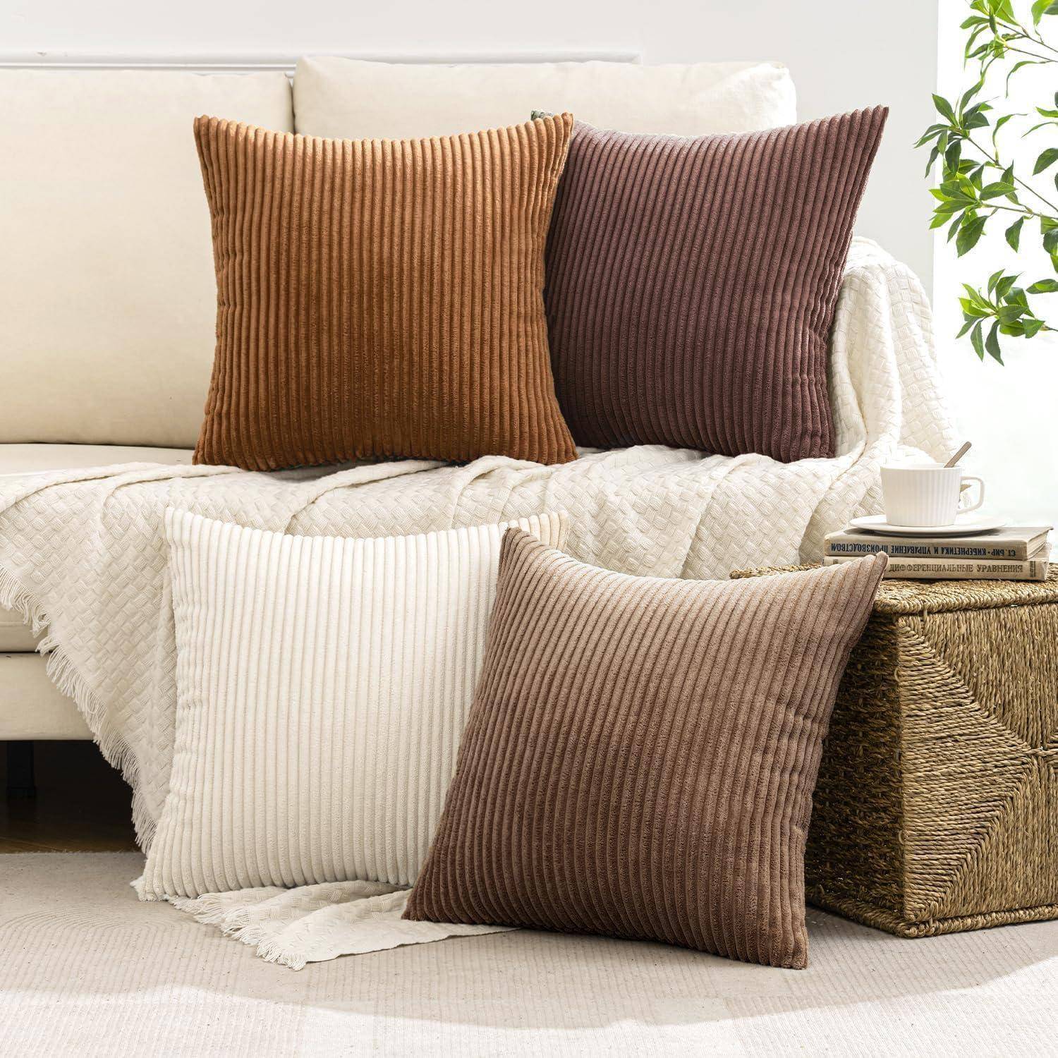 Vier gerippte Kissen in Braun-, Creme- und dunklen Schattierungen sind auf einem hellbeigen Sofa und einem Flechtkorb in einem gemütlichen Wohnbereich angeordnet.