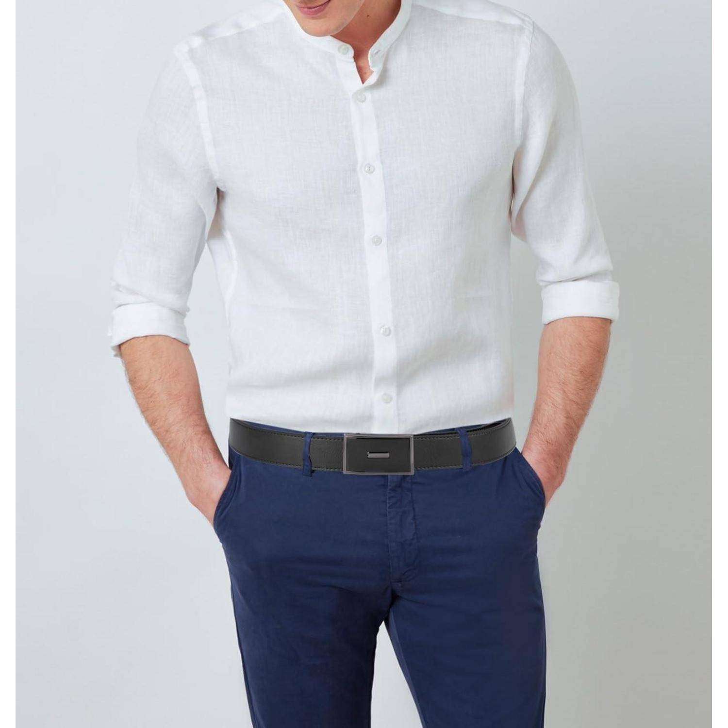 Eine Person, die ein weißes Hemd mit hochgekrempelten Ärmeln und eine dunkelblaue Hose trägt, steht mit in den Taschen verschränkten Händen vor einem neutralen Hintergrund.