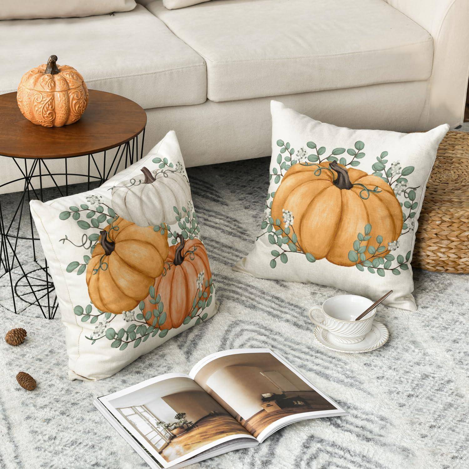 Kissen mit Kürbismotiven auf einem Teppich, in der Nähe eines Couchtisches mit einem kleinen Kürbis. Ein aufgeschlagenes Buch und eine Tasse sind neben dem Sofa. Gemütliche Atmosphäre.