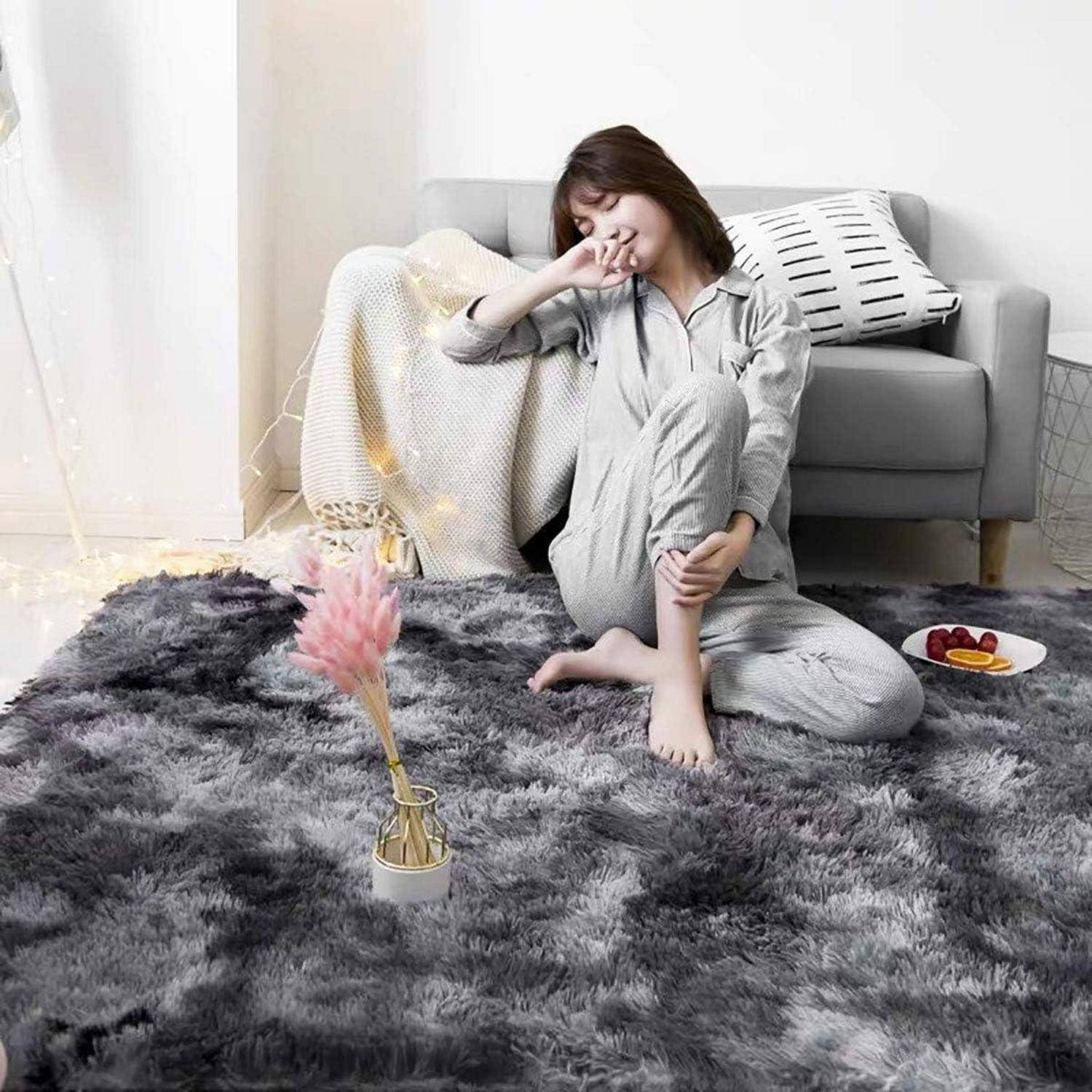 Eine Person in Pyjamas sitzt auf einem flauschigen grauen Teppich, gähnt und ist neben einem Sofa mit Kissen platziert. Ein kleiner Tisch mit Blumen und Snacks steht in der Nähe.