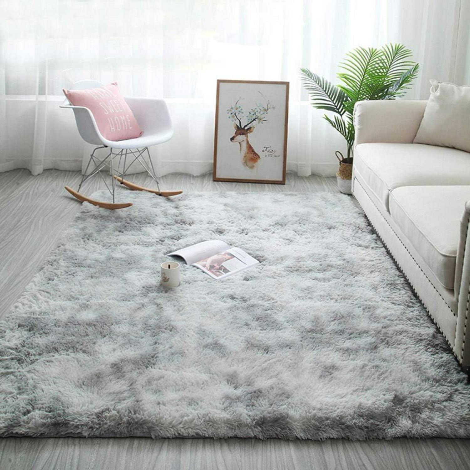 Ein gemütliches Wohnzimmer mit einem flauschigen grauen Teppich, einem weißen Schaukelstuhl mit rosa Kissen, Wandkunst mit einem Hirsch und einer Topfpflanze in der Nähe des Sofas.