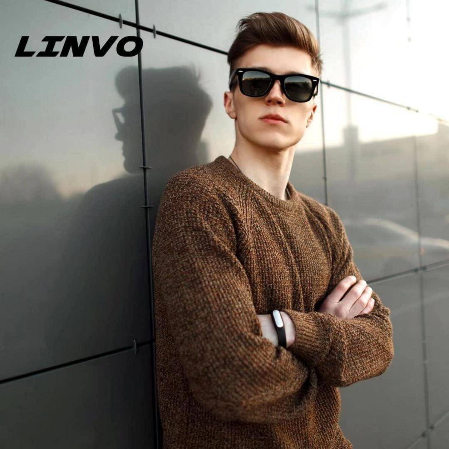 Eine Person mit Sonnenbrille und einem braunen Pullover steht selbstbewusst im Freien mit verschränkten Armen und zeigt eine modische Ausstrahlung.